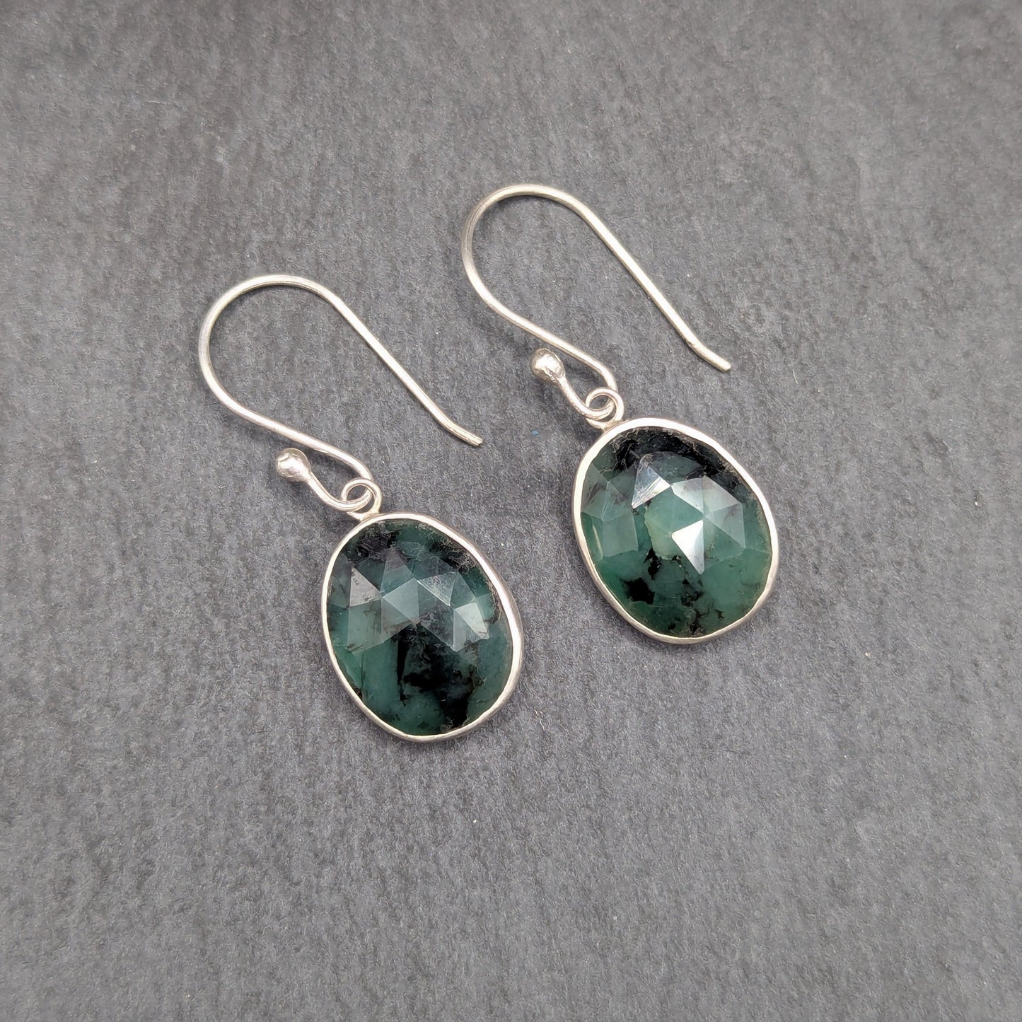 A pair of silver drop earrings with faceted dark green gemstones, displayed on a textured gray slate surface.