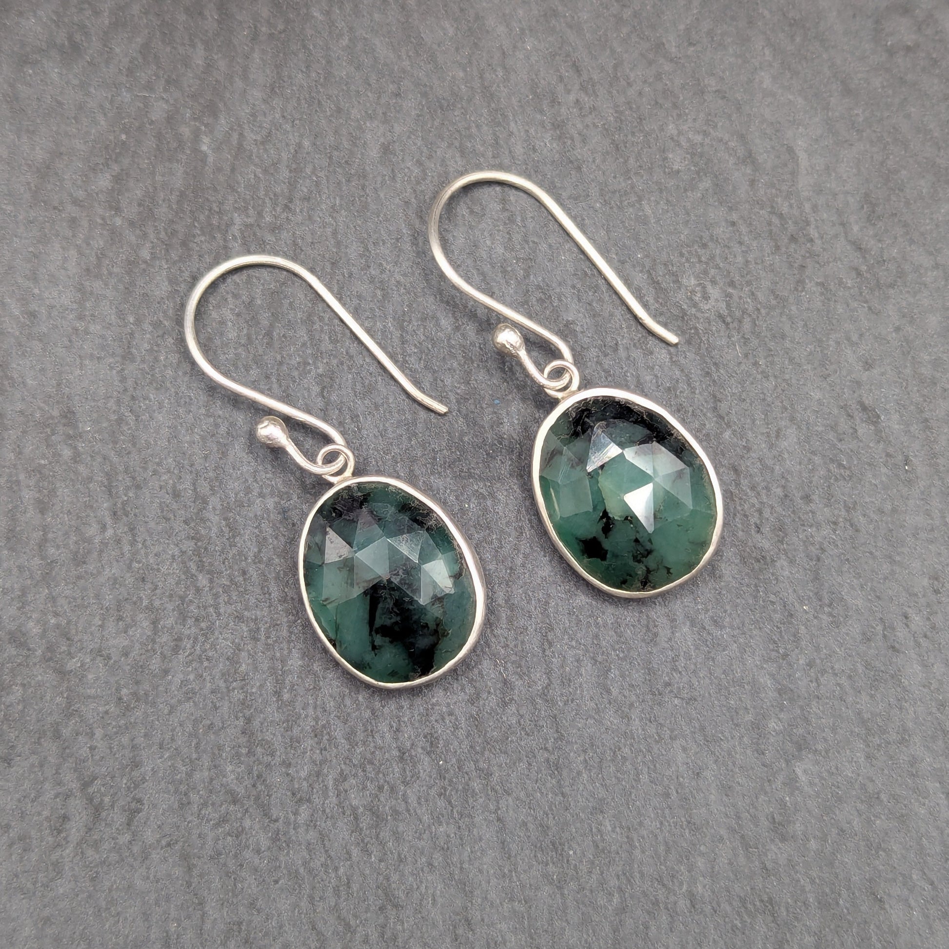 A pair of silver drop earrings with faceted dark green gemstones, displayed on a textured gray slate surface.