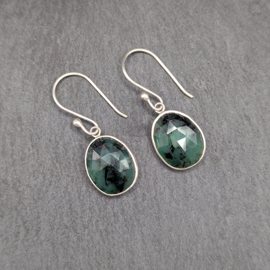 A pair of silver drop earrings with faceted dark green gemstones, displayed on a textured gray slate surface.